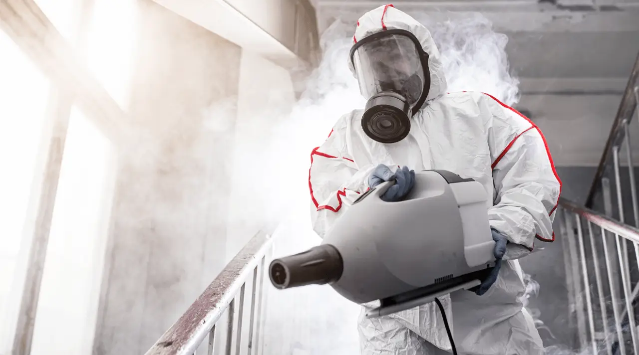 Biohazard cleanup worker disinfecting property with fogging machine in protective suit